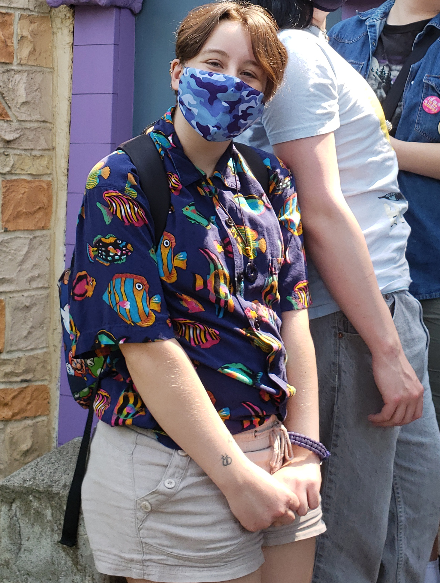 A photo of me, an androgynous white person with autistic body language and a fun tropical fish buttonup, smiling visibly even under a mask.
